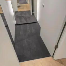 Położona ciemna wykładzina dywanowa w korytarzu, widoczna na podłodze pomiędzy drzwiami i fragmentem podłogi drewnianej. Widok z góry.