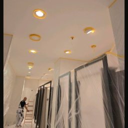 ML - Wnętrze w trakcie malowania: mężczyzna na drabinie, przygotowane ściany i sufit z oświetleniem, zabezpieczone drzwi folią malarską, taśmy ochronne na krawędziach.