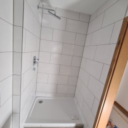 ML - Wykończona kabina prysznicowa z białymi, prostokątnymi płytkami ułożonymi poziomo, chromowaną armaturą i brodzikiem. Częściowo widoczna toaleta i akcesoria glazurnicze.