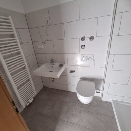 ML - Wykończona łazienka z białymi, prostokątnymi płytkami na ścianach i szarymi płytkami na podłodze. Widoczna umywalka, toaleta i grzejnik drabinkowy.