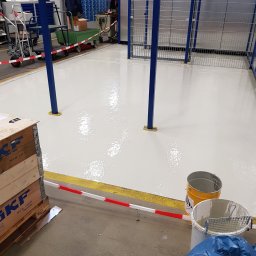 ML - Świeżo pomalowana, lśniąca podłoga w hali magazynowej z niebieskimi słupami i metalową siatką. Obok stoją puszki z farbą i skrzynia transportowa SKF.