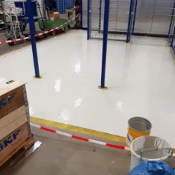 Świeżo pomalowana, lśniąca podłoga w hali magazynowej z niebieskimi słupami i metalową siatką. Obok stoją puszki z farbą i skrzynia transportowa SKF.