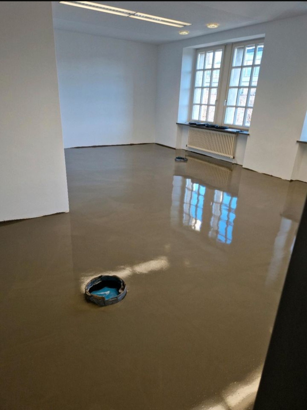 Wnętrze po wylaniu samopoziomującej wylewki. Widoczne otwory instalacyjne i refleksy okien w gładkiej powierzchni podłogi. Minimalistyczny, nowoczesny styl.