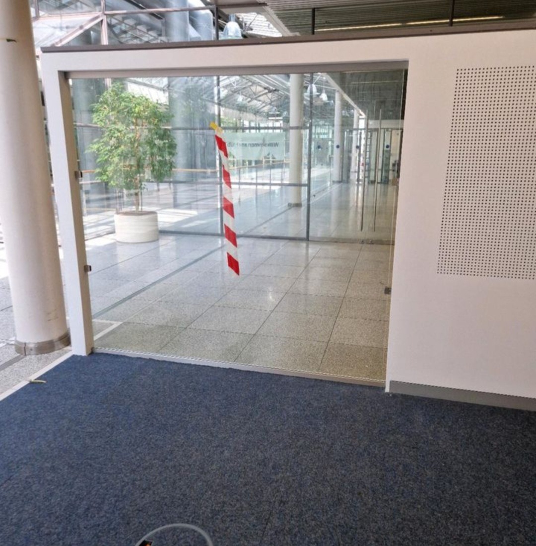 Szklane drzwi przesuwne z czerwoną taśmą ostrzegawczą w nowoczesnym budynku z roślinami i kolumnami. Widok z wnętrza na przeszkloną przestrzeń.