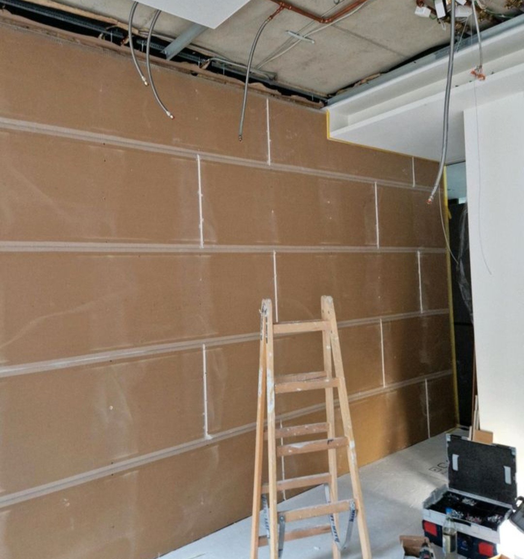 Montaż płyt kartonowo-gipsowych na ścianie z widoczną drabiną i instalacją elektryczną pod sufitem. Etap wykończeniowy w pomieszczeniu.