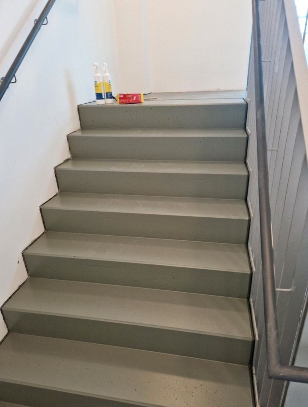 Pomalowane schody wewnątrz budynku, widoczne środki czyszczące i narzędzia malarskie na górnym stopniu. Ujęcie z dołu, skupione na wykończeniu.