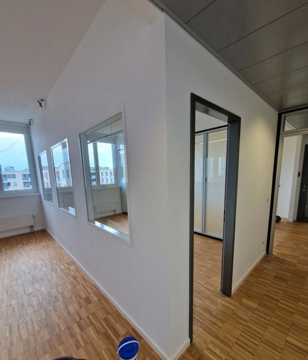 Wnętrze biura po malowaniu: białe ściany, drewniana podłoga, okna z widokiem na miasto. Widoczny fragment puszki z farbą.