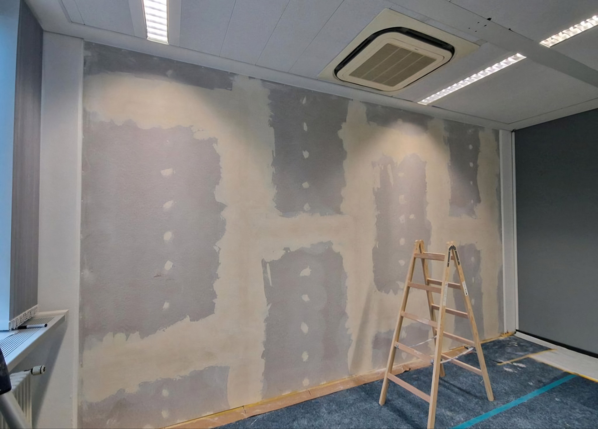 Przygotowanie ściany do malowania: szpachlowanie i gruntowanie. Drabina stoi przy ścianie. Widoczne ślady po łataniu ubytków. Oświetlenie sufitowe.