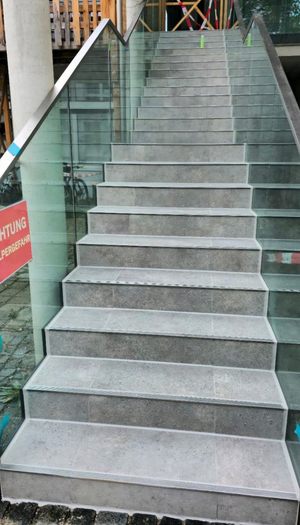 Wykończone schody z szarych płytek z subtelnym wzorem, zabezpieczone szklaną balustradą z metalowymi poręczami. Widok z dołu, lekko z boku.