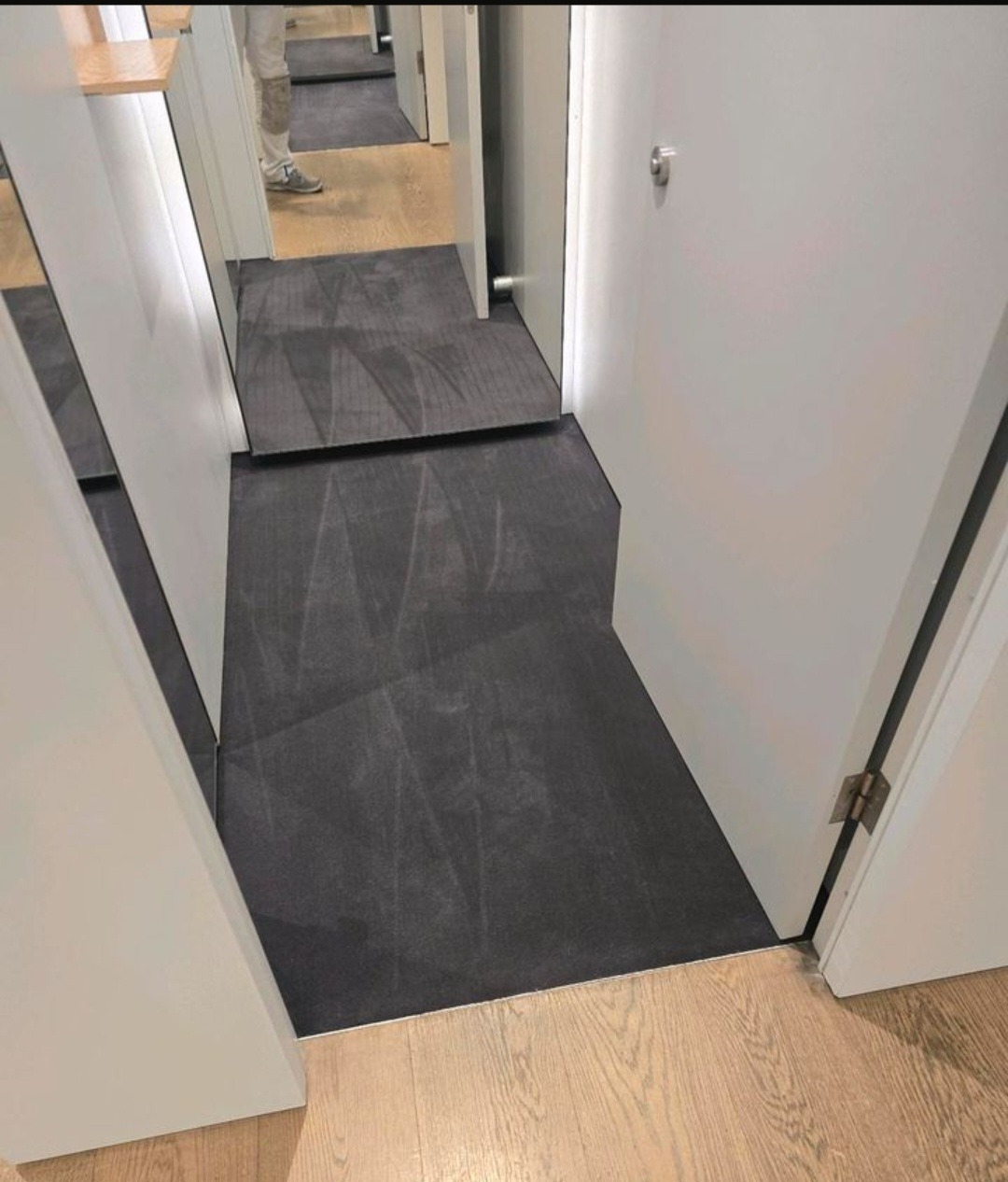 Położona ciemna wykładzina dywanowa w korytarzu, widoczna na podłodze pomiędzy drzwiami i fragmentem podłogi drewnianej. Widok z góry.