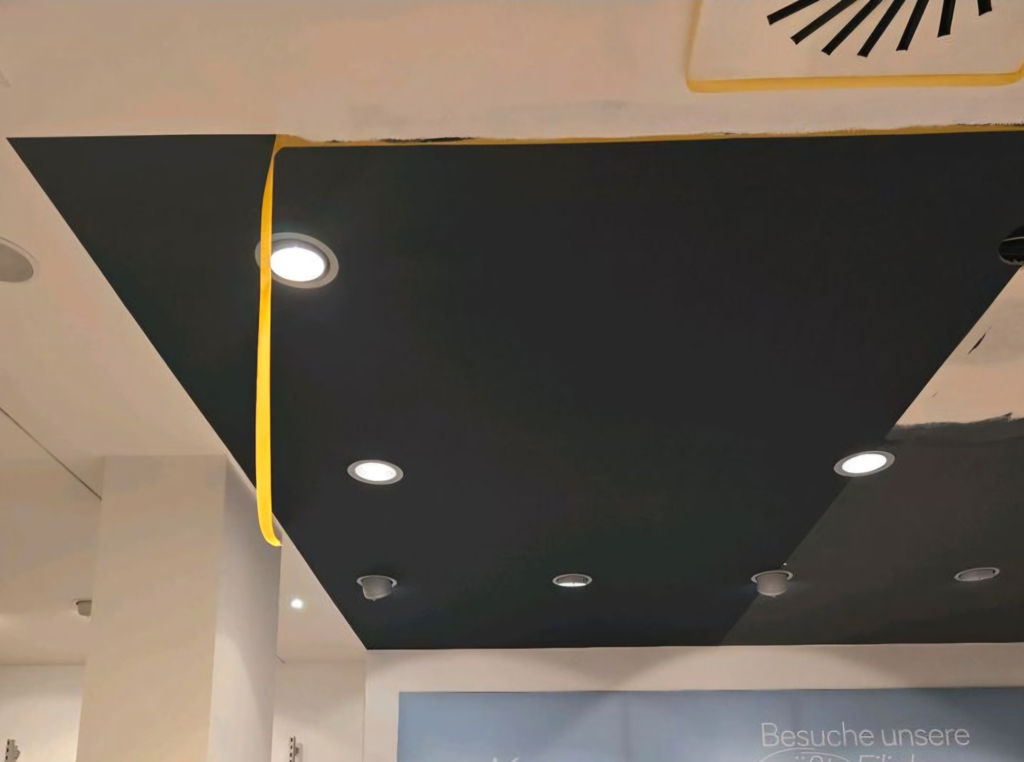 Czarne wykończenie sufitu z wbudowanymi lampami, częściowo zakryte taśmą malarską, w trakcie prac wykończeniowych. Widoczny fragment wentylacji i ściana.