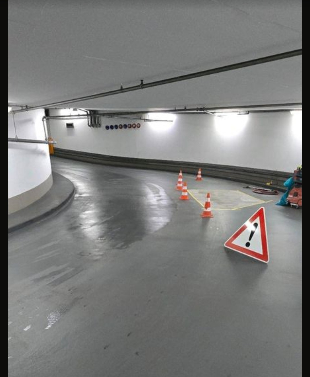 Świeżo pomalowana posadzka w garażu podziemnym, z widocznymi stożkami ostrzegawczymi i znakiem trójkątnym. Powierzchnia mokra, odbijająca światło.