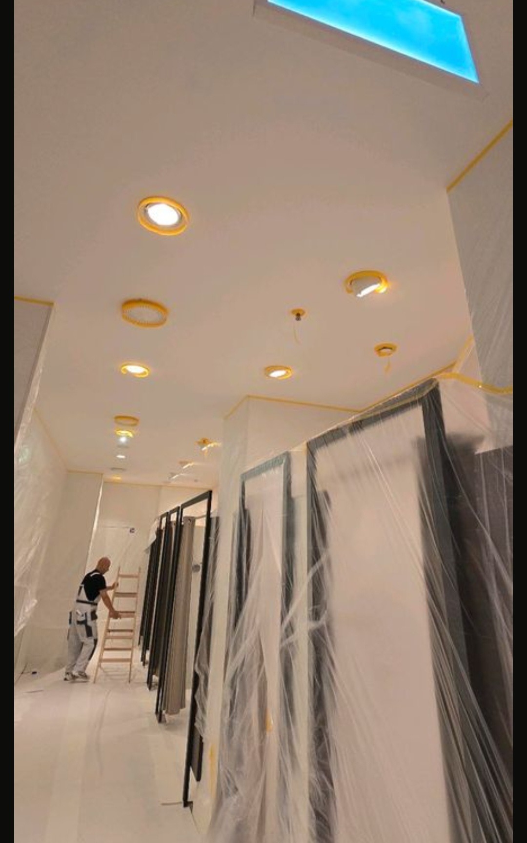 Wnętrze w trakcie malowania: mężczyzna na drabinie, przygotowane ściany i sufit z oświetleniem, zabezpieczone drzwi folią malarską, taśmy ochronne na krawędziach.