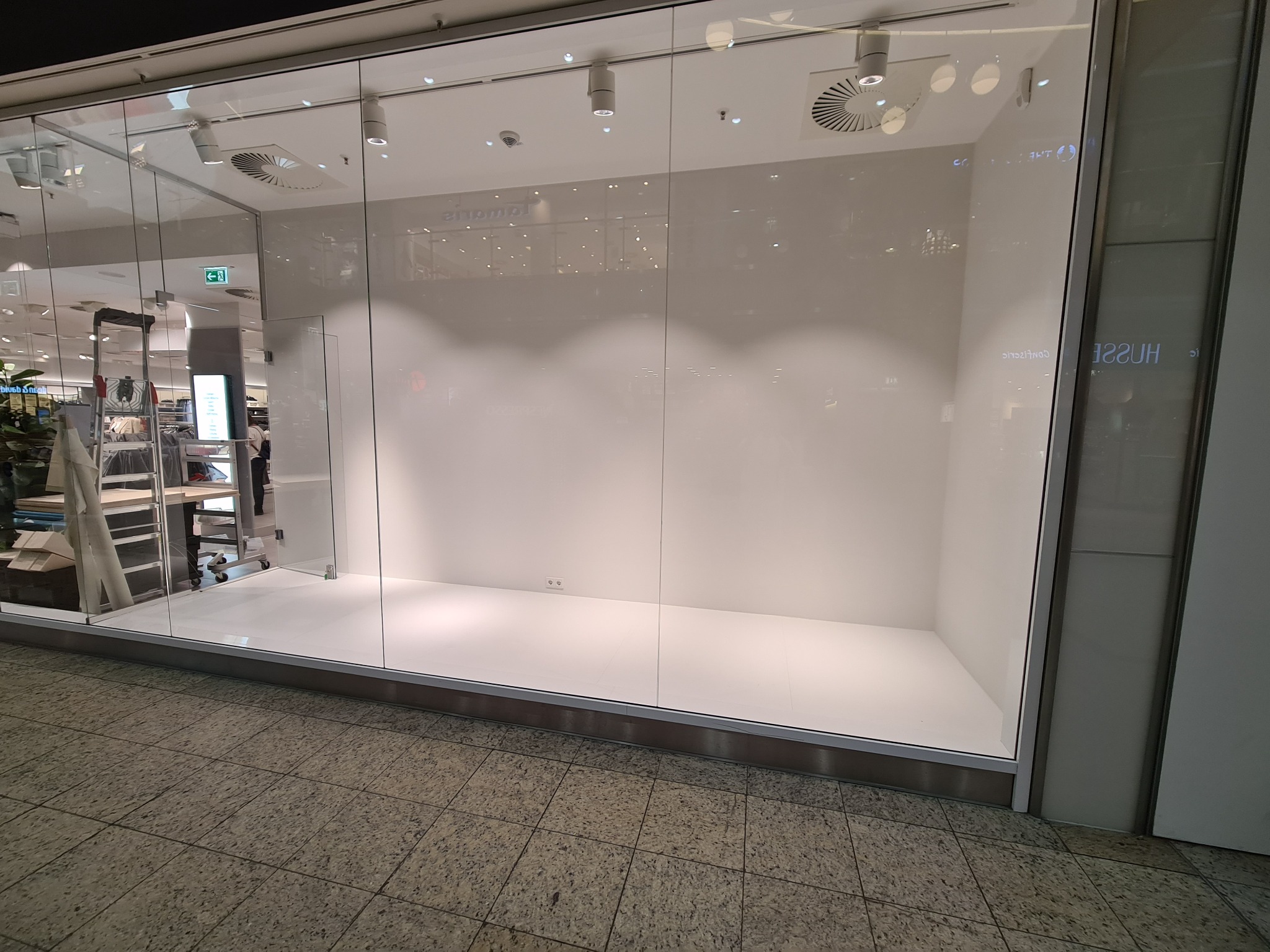 Pusta, oświetlona witryna sklepowa z białymi ścianami i podłogą, widoczna z zewnątrz przez szklaną fasadę. W tle widoczne wnętrze centrum handlowego.