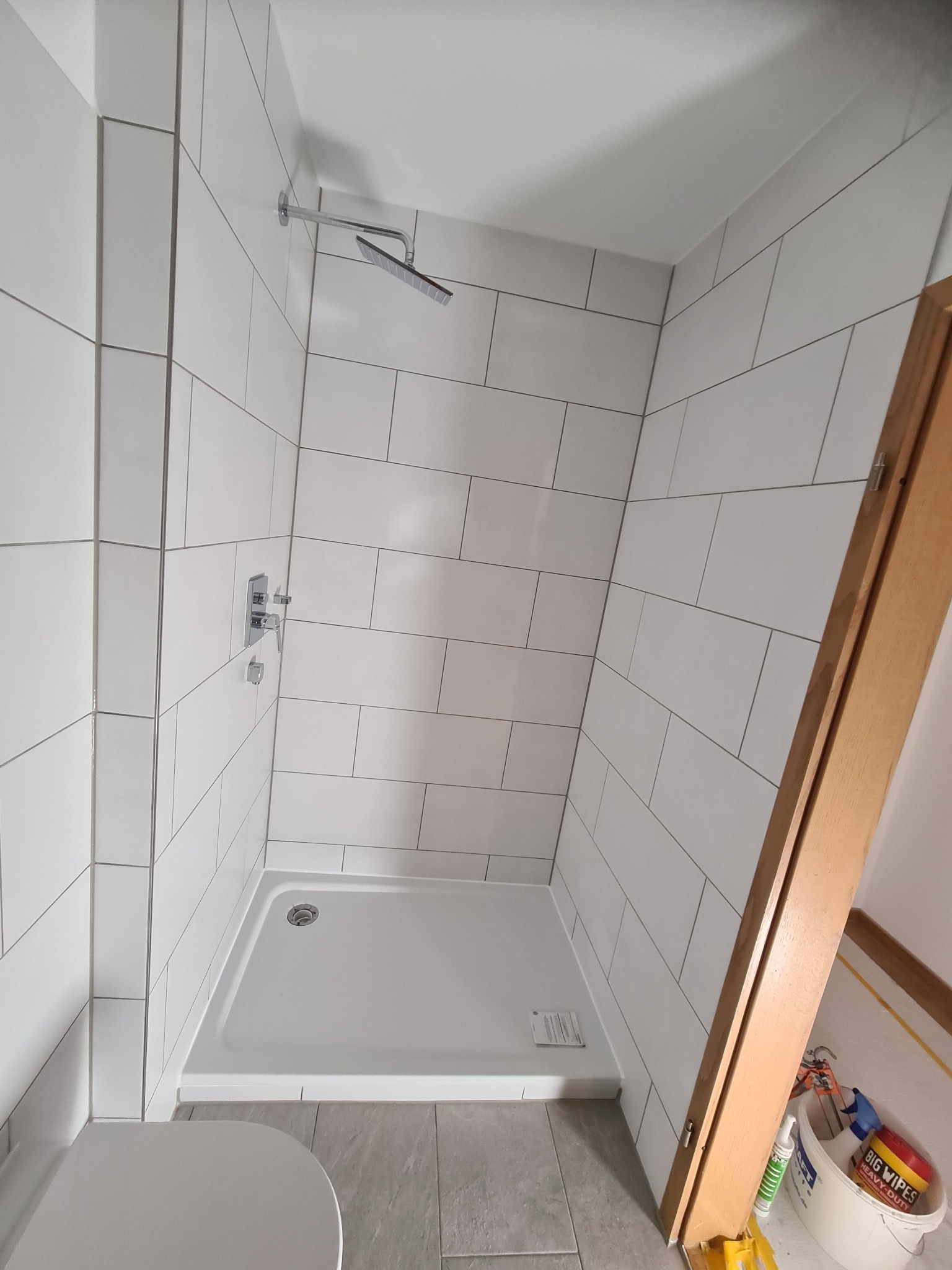 Wykończona kabina prysznicowa z białymi, prostokątnymi płytkami ułożonymi poziomo, chromowaną armaturą i brodzikiem. Częściowo widoczna toaleta i akcesoria glazurnicze.