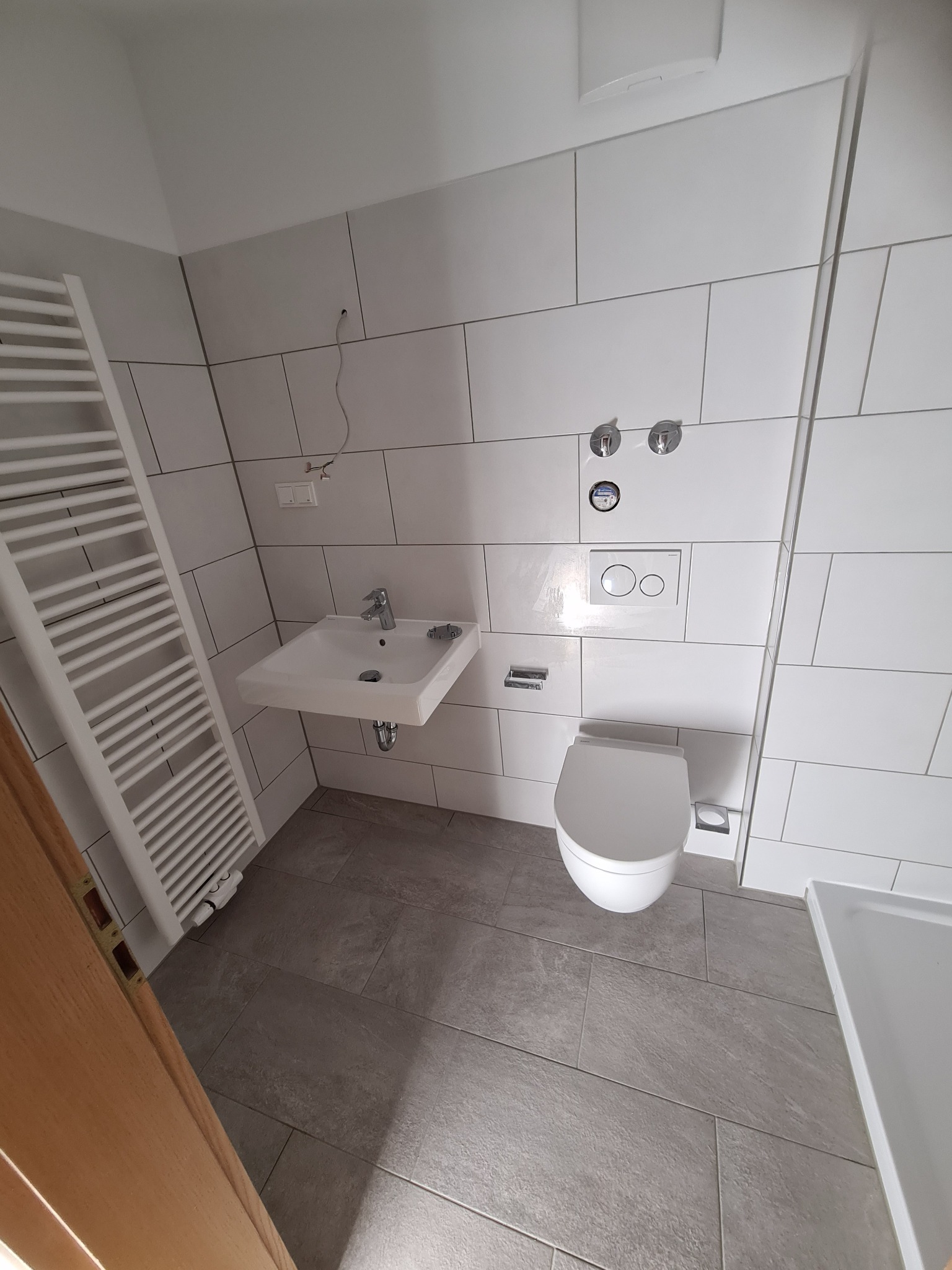 Wykończona łazienka z białymi, prostokątnymi płytkami na ścianach i szarymi płytkami na podłodze. Widoczna umywalka, toaleta i grzejnik drabinkowy.