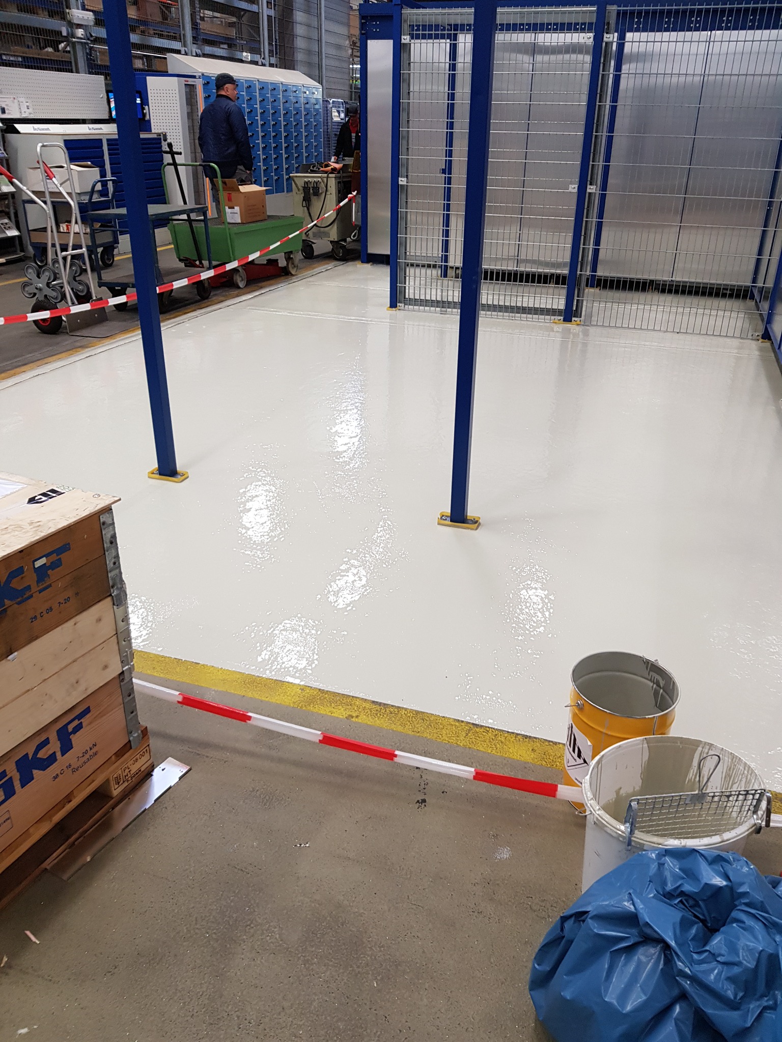 Świeżo pomalowana, lśniąca podłoga w hali magazynowej z niebieskimi słupami i metalową siatką. Obok stoją puszki z farbą i skrzynia transportowa SKF.