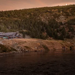 Schronisko górskie z oświetlonymi oknami na tle zalesionego zbocza i częściowo zamarzniętego jeziora, pod pomarańczowym niebem z widocznym księżycem.