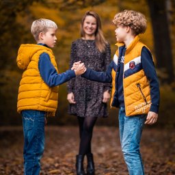 Patryk Michalski fotografia i grafika - Jesienna sesja rodzinna: Dwóch chłopców w żółtych kamizelkach ściska dłonie na tle uśmiechniętej kobiety w parku, opadłe liście, ciepłe światło.