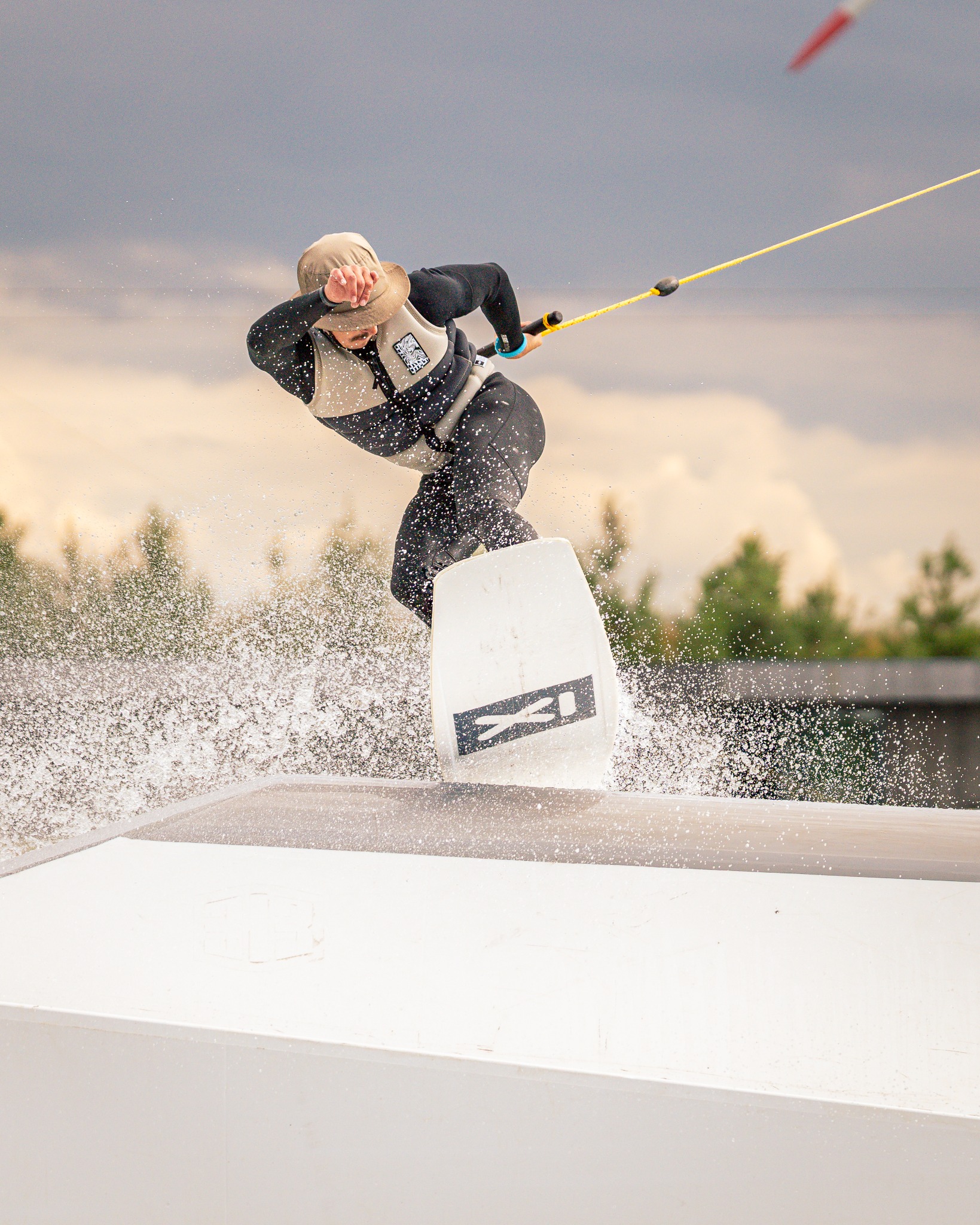 Wakeboardzista w kapeluszu wykonuje trik na rampie, z fontanną wody i pochmurnym niebem w tle. Dynamiczne ujęcie z bliska, podkreślające akcję i sportowy charakter.