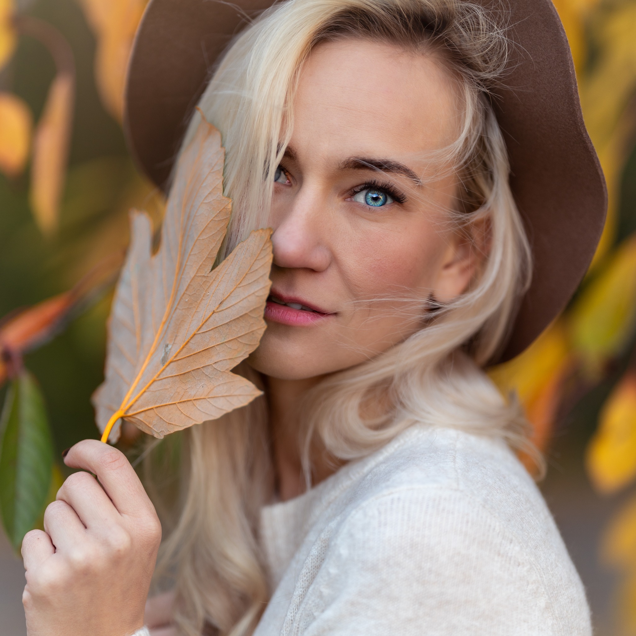 Portret kobiety z blond włosami w brązowym kapeluszu, trzymającej jesienny liść na tle rozmytych żółtych i zielonych liści. Uwagę przyciągają jej intensywnie niebieskie oczy.