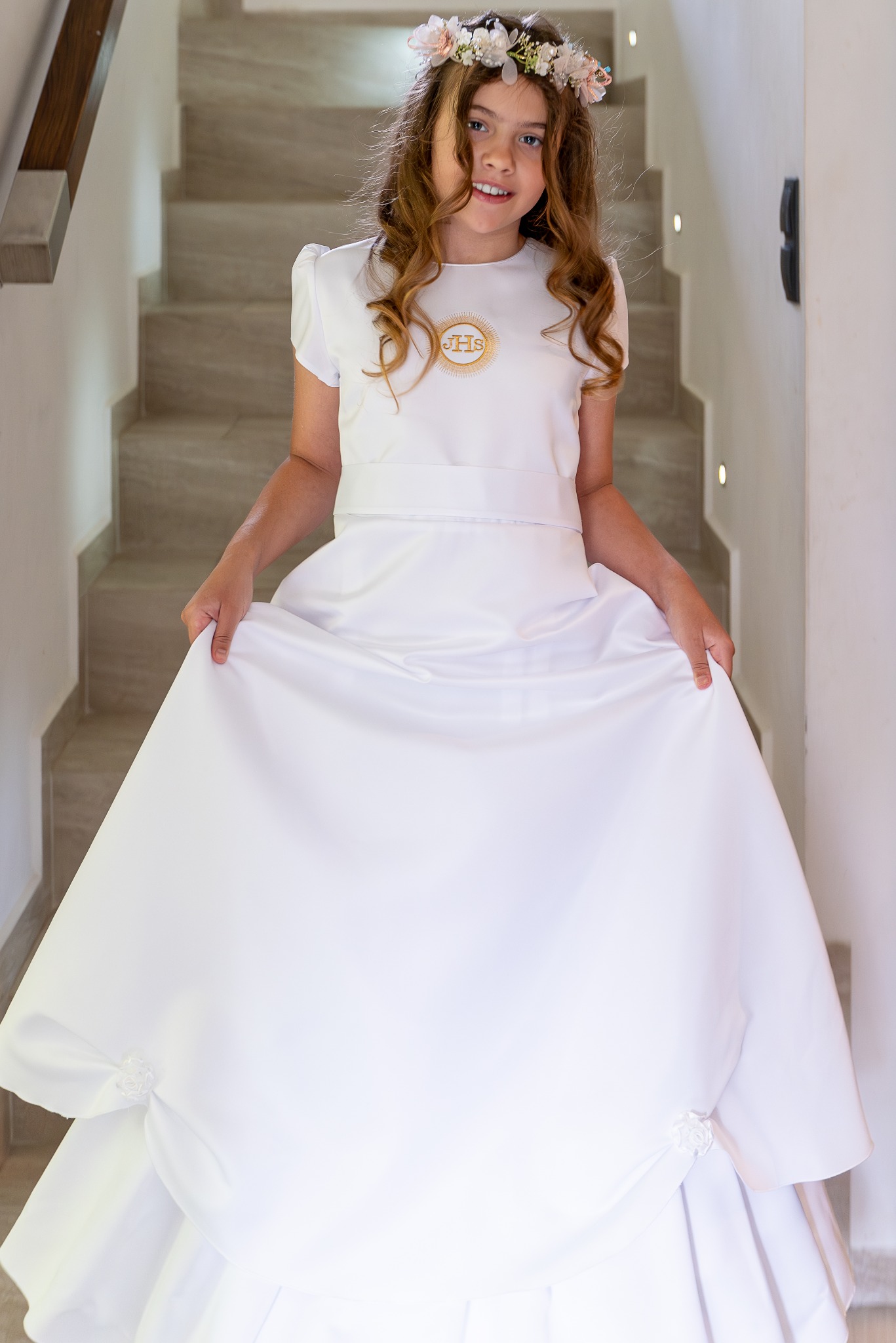 Dziewczynka w białej sukience komunijnej z wiankiem na głowie, pozująca na tle schodów. Sukienka zdobiona symbolem IHS. Uśmiechnięta, naturalne światło.