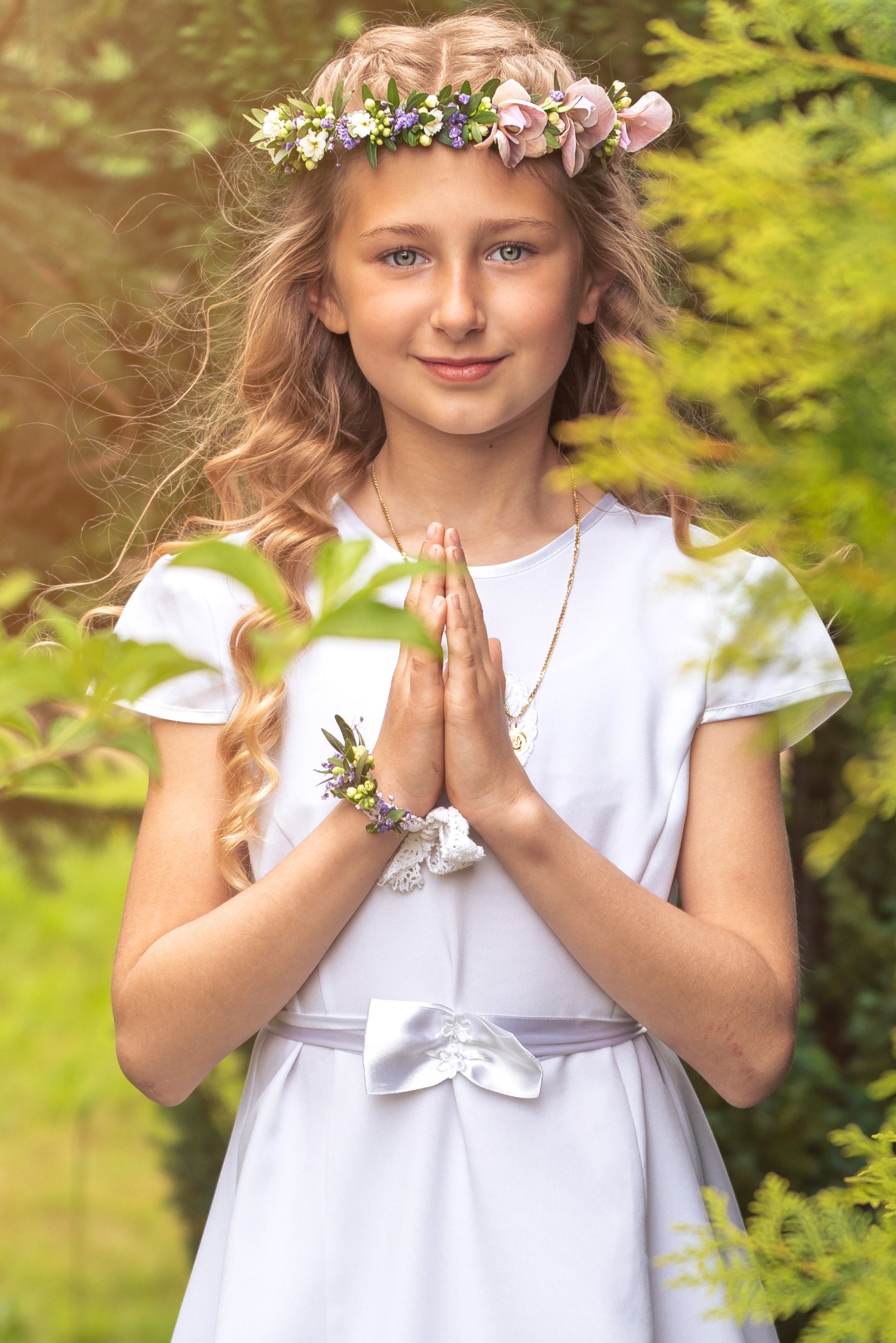 Portret dziewczynki w białej sukience komunijnej z wiankiem na głowie, dłonie złożone do modlitwy, na tle zieleni. Delikatne światło, ciepła kolorystyka, naturalny wygląd.
