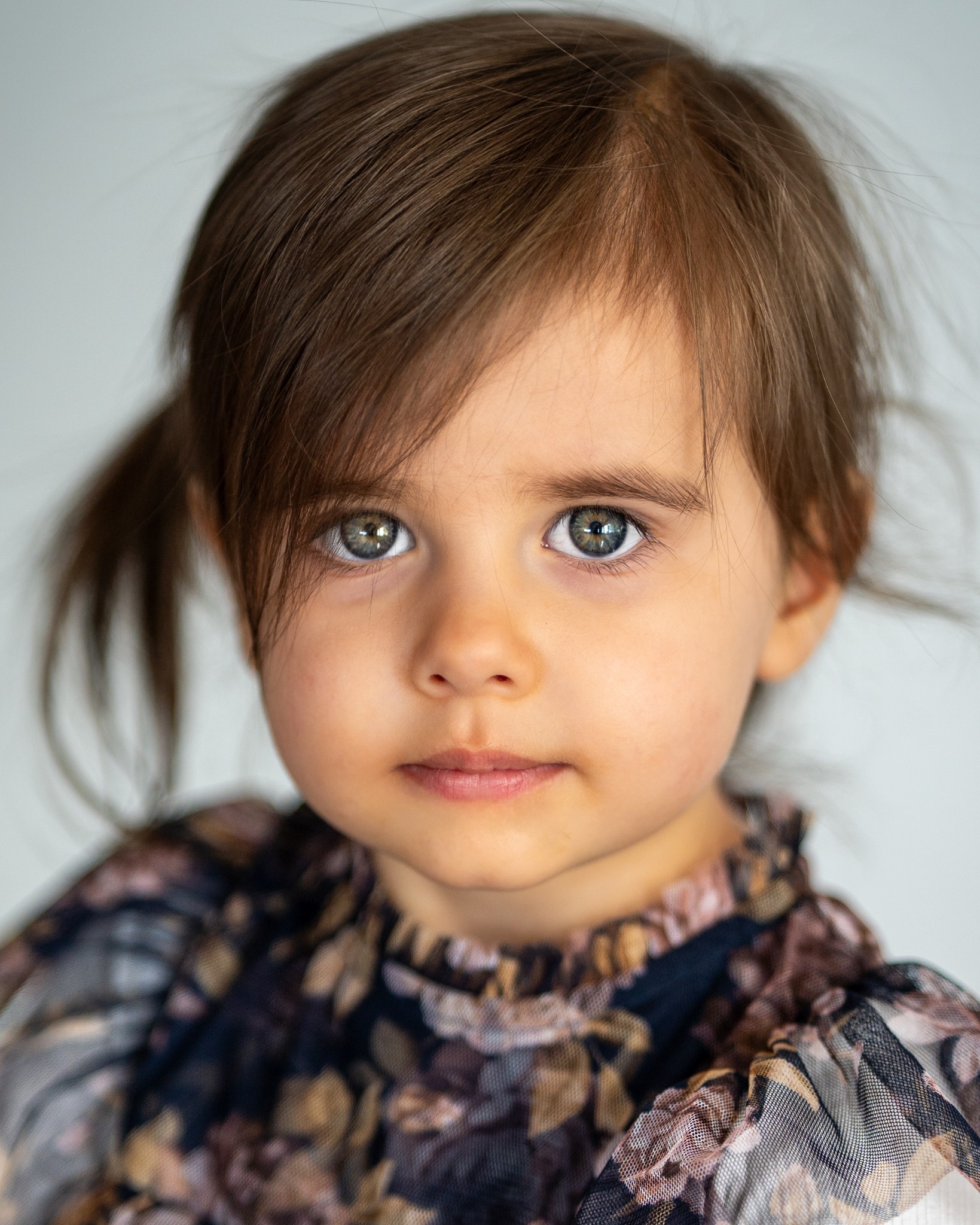 Portret uroczej dziewczynki z brązowymi włosami i zielonymi oczami, ubrana w bluzkę w kwiaty. Delikatny uśmiech i naturalne światło podkreślają jej dziecięcy wdzięk.