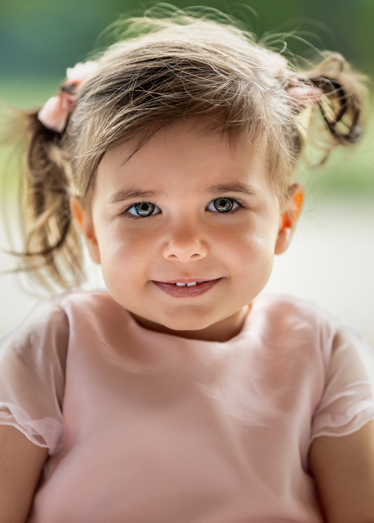 Portret uśmiechniętej dziewczynki z brązowymi włosami spiętymi w dwa kucyki z różowymi kokardkami, na rozmytym, zielonym tle.