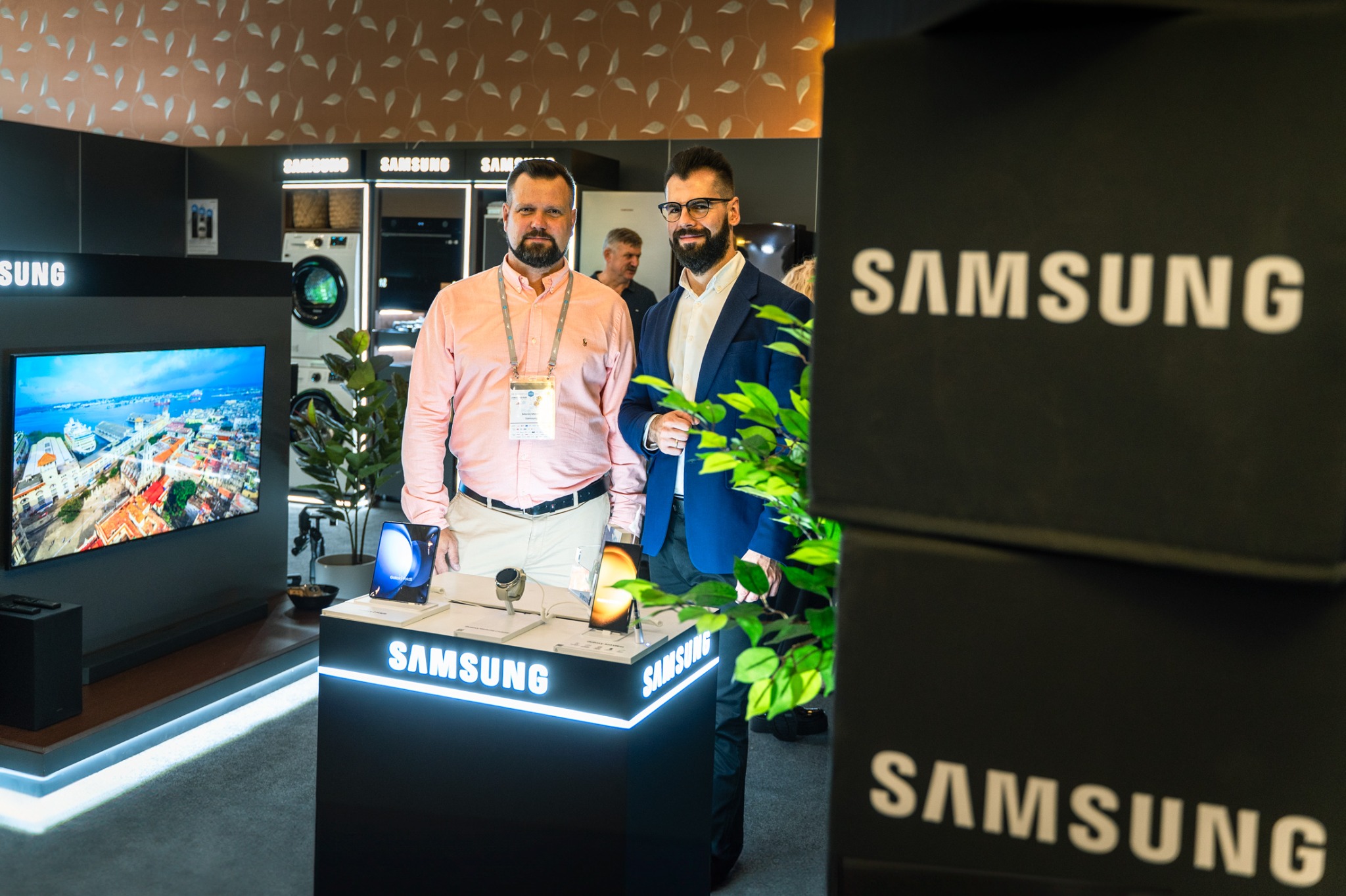 Dwóch mężczyzn na tle stoiska Samsung z telewizorem, pralką i smartfonami. Ekspozycja oświetlona neonowym światłem, w tle widoczni inni ludzie.