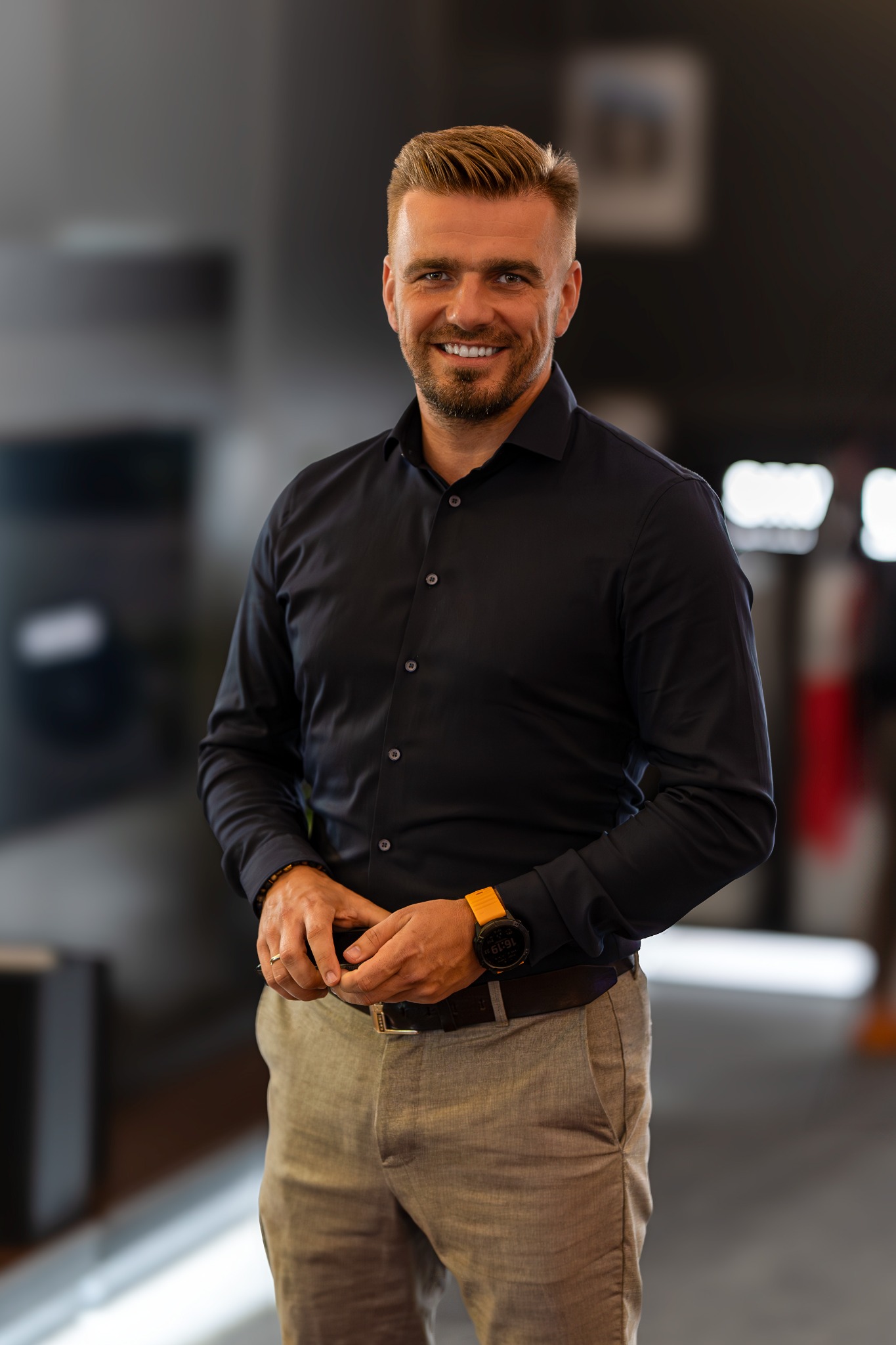 Uśmiechnięty mężczyzna w czarnej koszuli i beżowych spodniach, z zegarkiem na ręku. Portret biznesowy w neutralnym wnętrzu, z delikatnym rozmyciem tła.