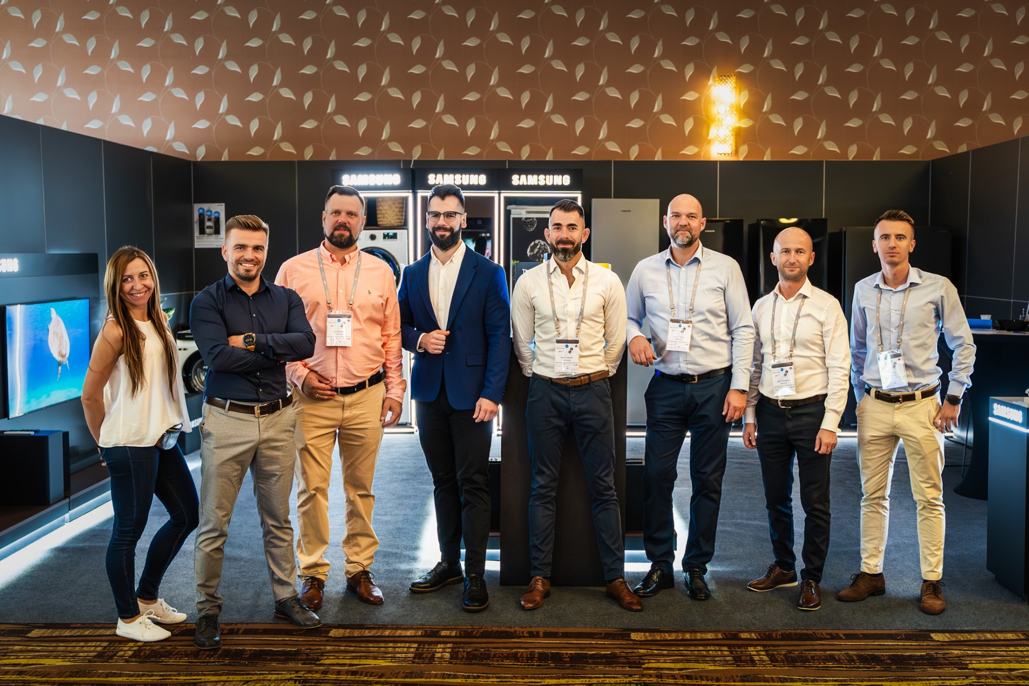 Grupa osób (mężczyzn i kobieta) w eleganckich strojach, stojących na tle ekspozycji sprzętu AGD Samsung, z identyfikatorami na szyjach, pozujących do zdjęcia.