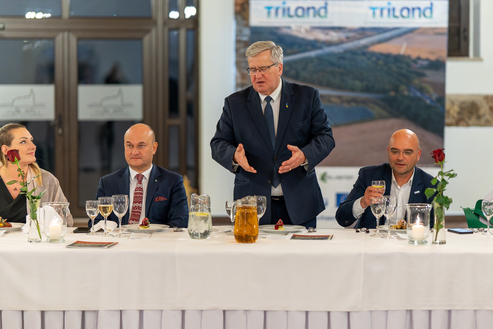 Mężczyzna w garniturze przemawia na tle baneru Trilando podczas uroczystej kolacji, z gośćmi i kwiatami na stole.