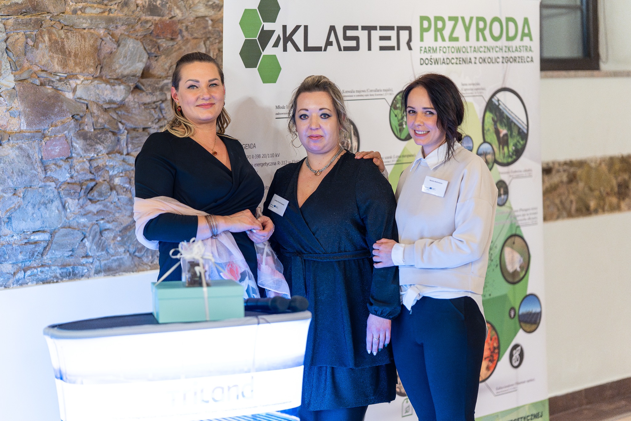 Trzy kobiety pozują na tle kamiennej ściany i baneru informacyjnego 'Klaster Przyroda' na evencie; widoczny stół z prezentem.