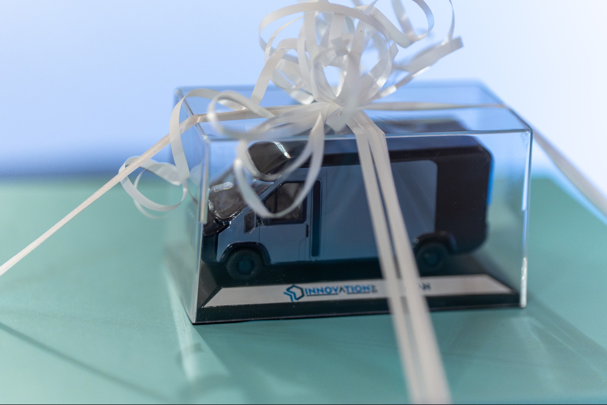 Model furgonetki w przezroczystym pudełku, ozdobionym białą wstążką, leżący na turkusowym tle. Logo firmy 'Innovation' widoczne na podstawie modelu.