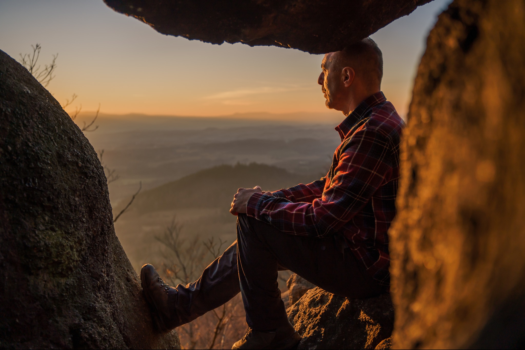 Mężczyzna w flanelowej koszuli siedzi na skale, kontemplując górski krajobraz o zmierzchu. Ciepłe światło podkreśla teksturę skał i odległe wzgórza.