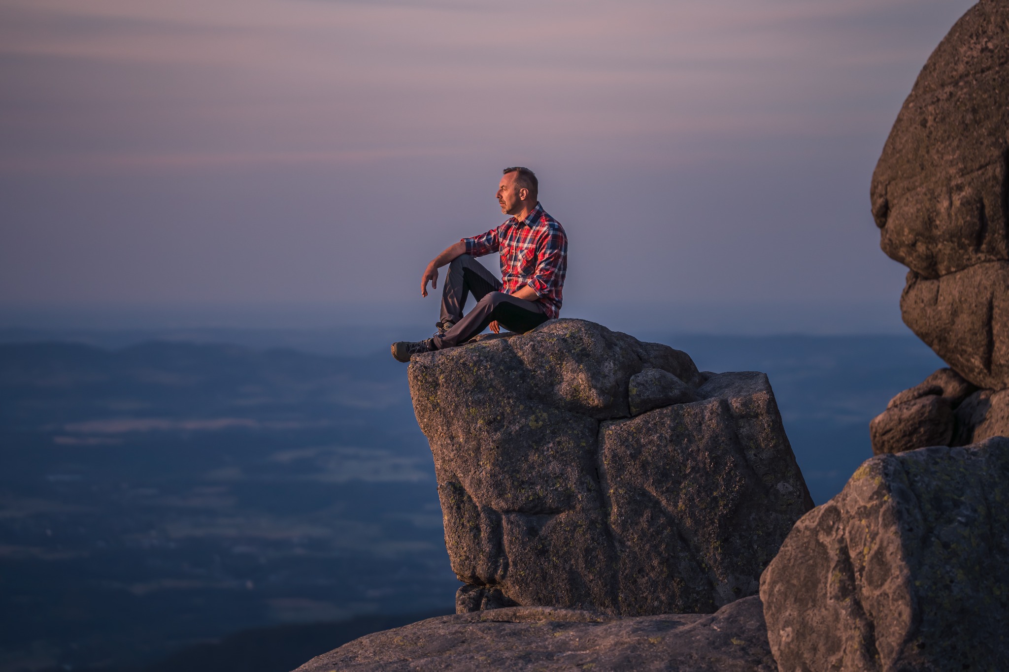 Mężczyzna w kraciastej koszuli siedzi na szczycie skalnej formacji z widokiem na rozległy krajobraz o zmierzchu, spokojna sceneria.
