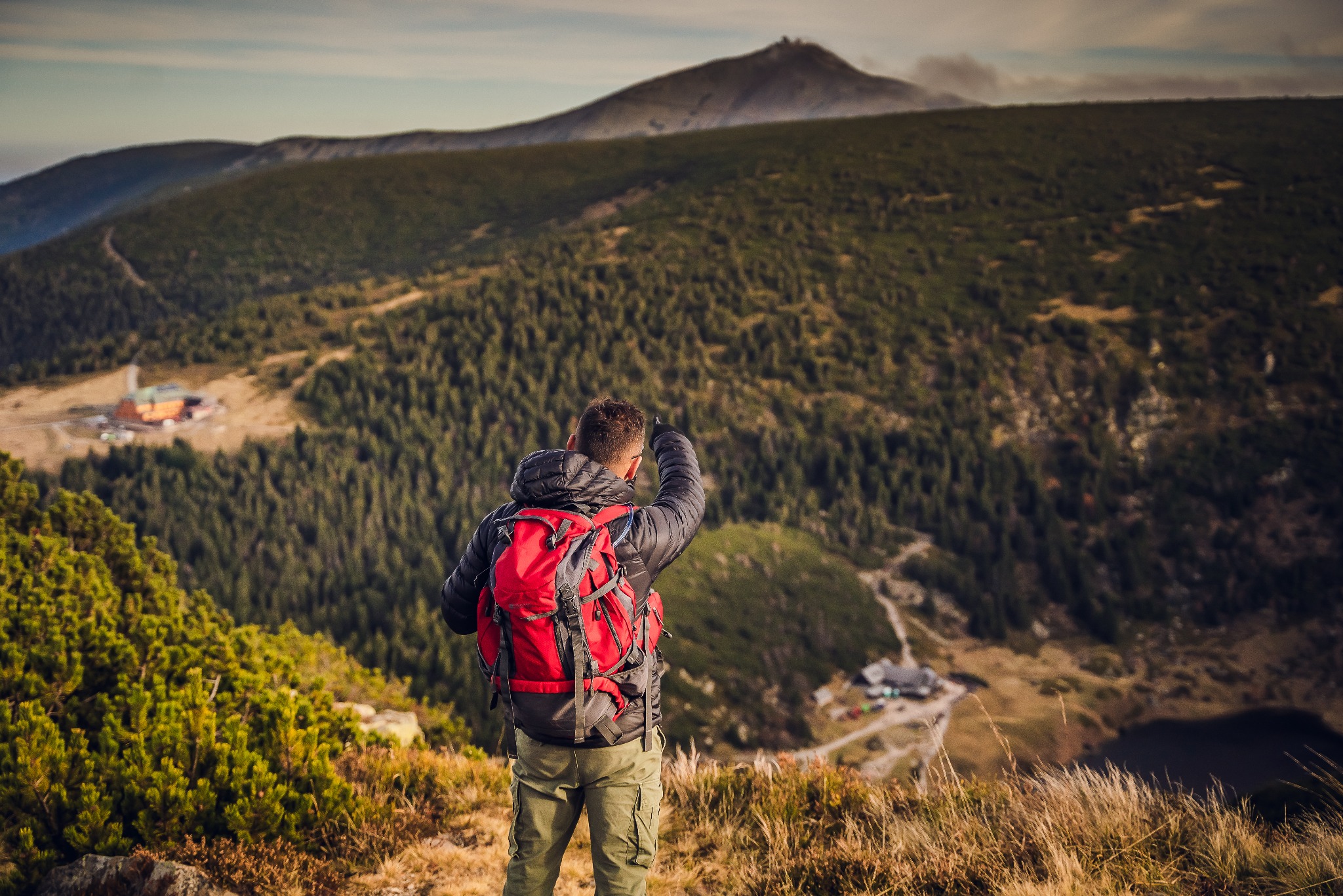 Turysta z czerwonym plecakiem podziwia górski krajobraz z widokiem na szczyt i schronisko. Mężczyzna w kurtce wskazuje na odległy punkt.