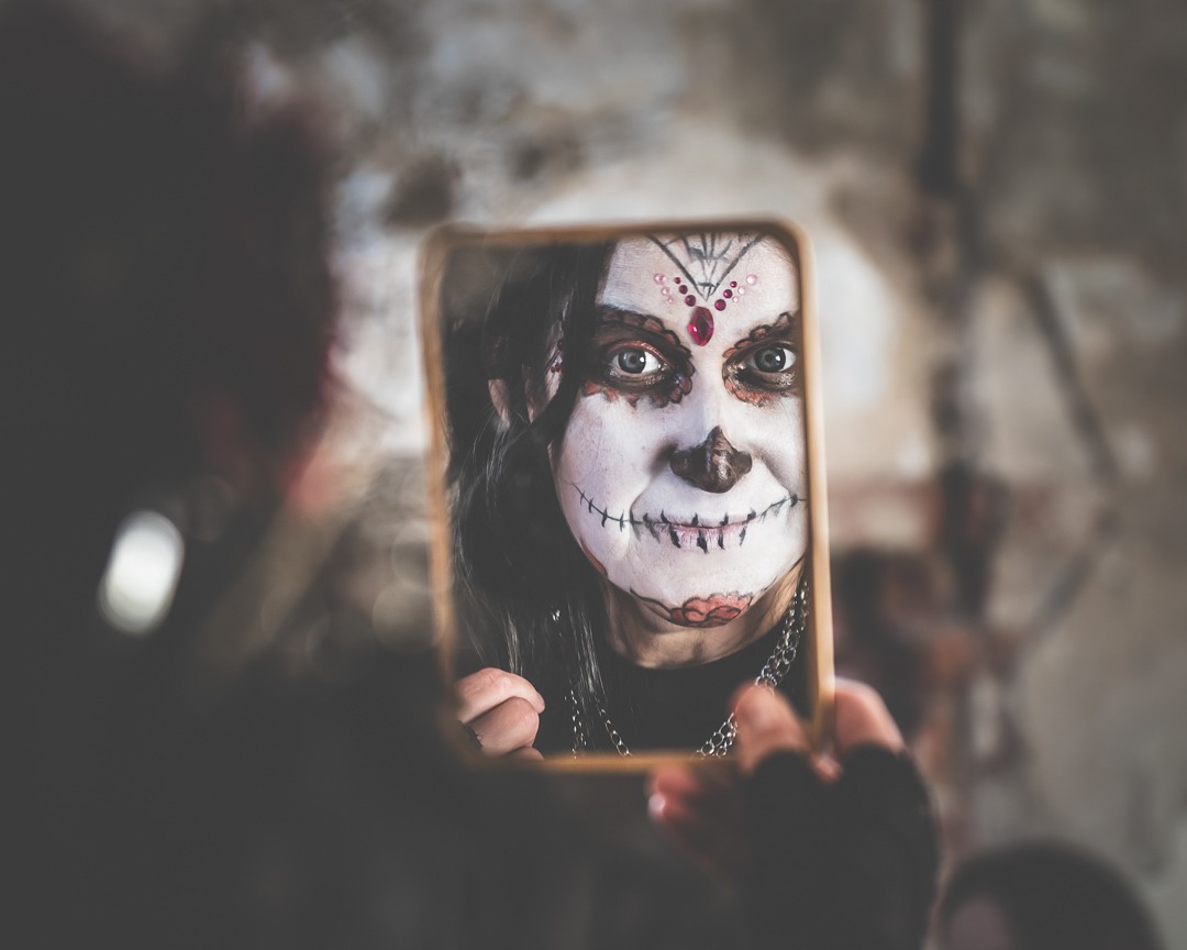 Portret kobiety z makijażem inspirowanym Día de Muertos, odbity w lustrze trzymanym w dłoniach, wykonany w Legnickim Polu. Mroczny klimat, artystyczny make-up.