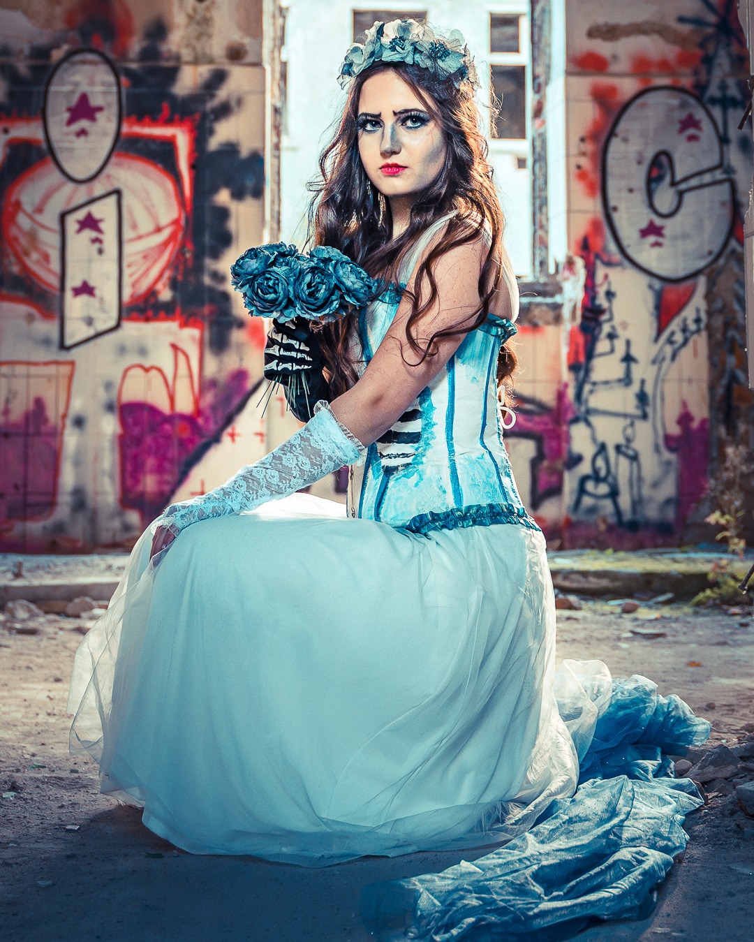 Kobieta w stylizowanej sukni ślubnej, z bukietem niebieskich róż, siedzi na tle ściany z graffiti w opuszczonym budynku. Gotycki makijaż i wianek na głowie.