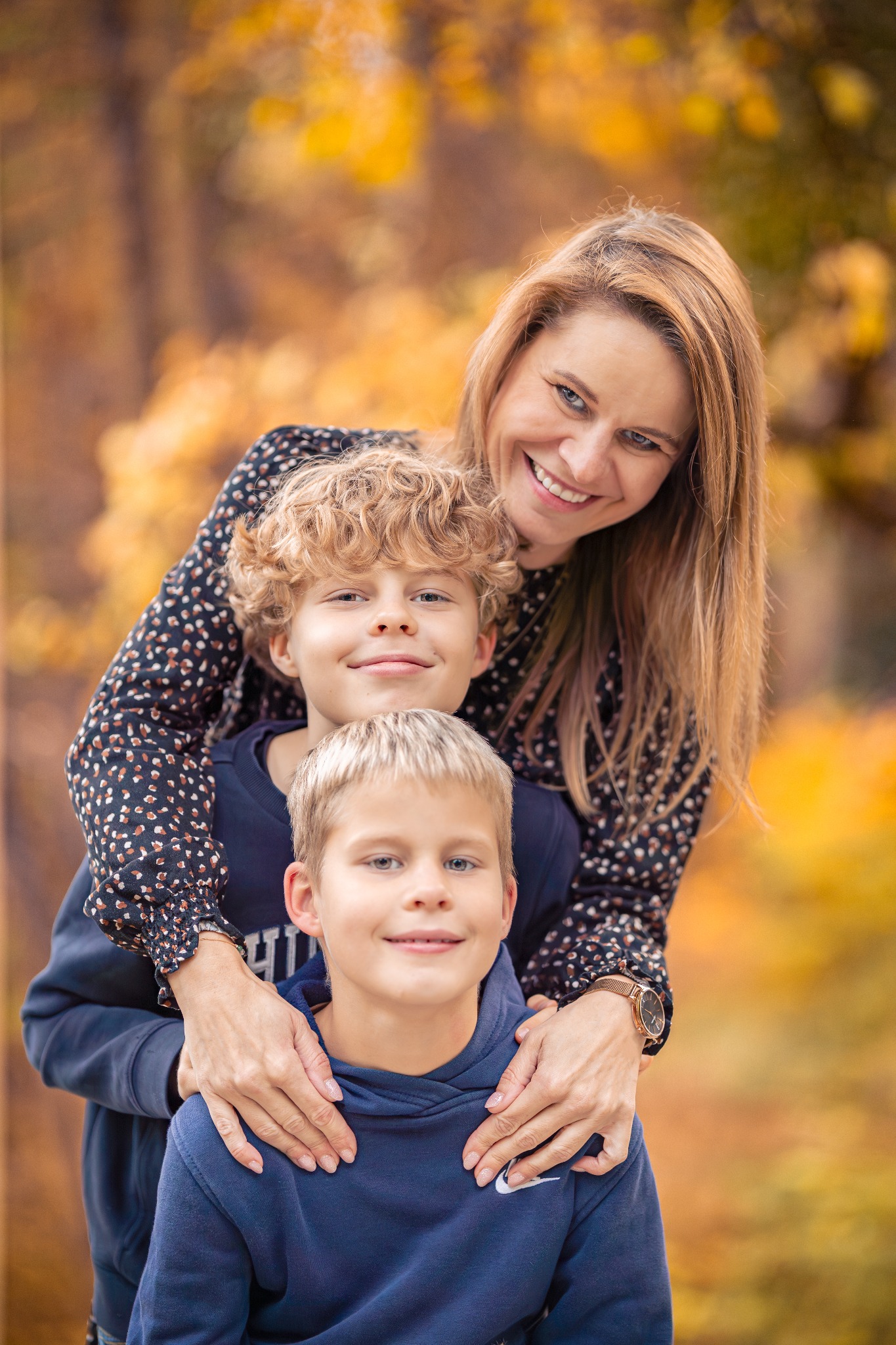 Portret rodzinny w jesiennej scenerii: uśmiechnięta mama obejmuje dwóch synów, z których jeden stoi przed drugim. Ciepłe, naturalne światło podkreśla bliskość.