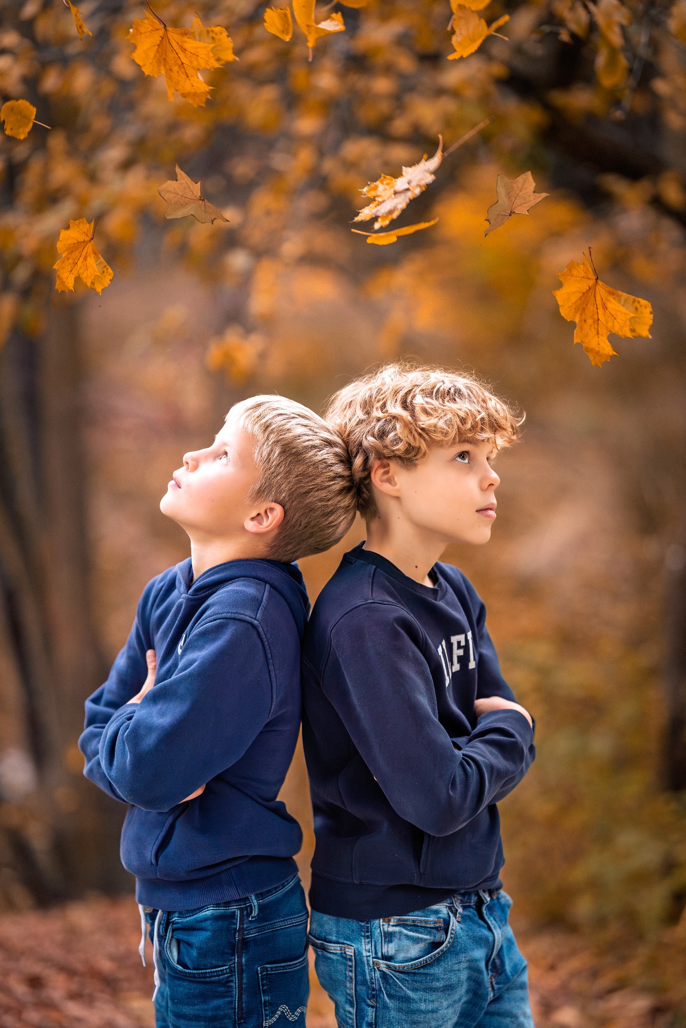 Jesienna sesja portretowa dwóch chłopców opartych plecami o siebie, spoglądających w górę na spadające liście klonu w ciepłych, jesiennych barwach.