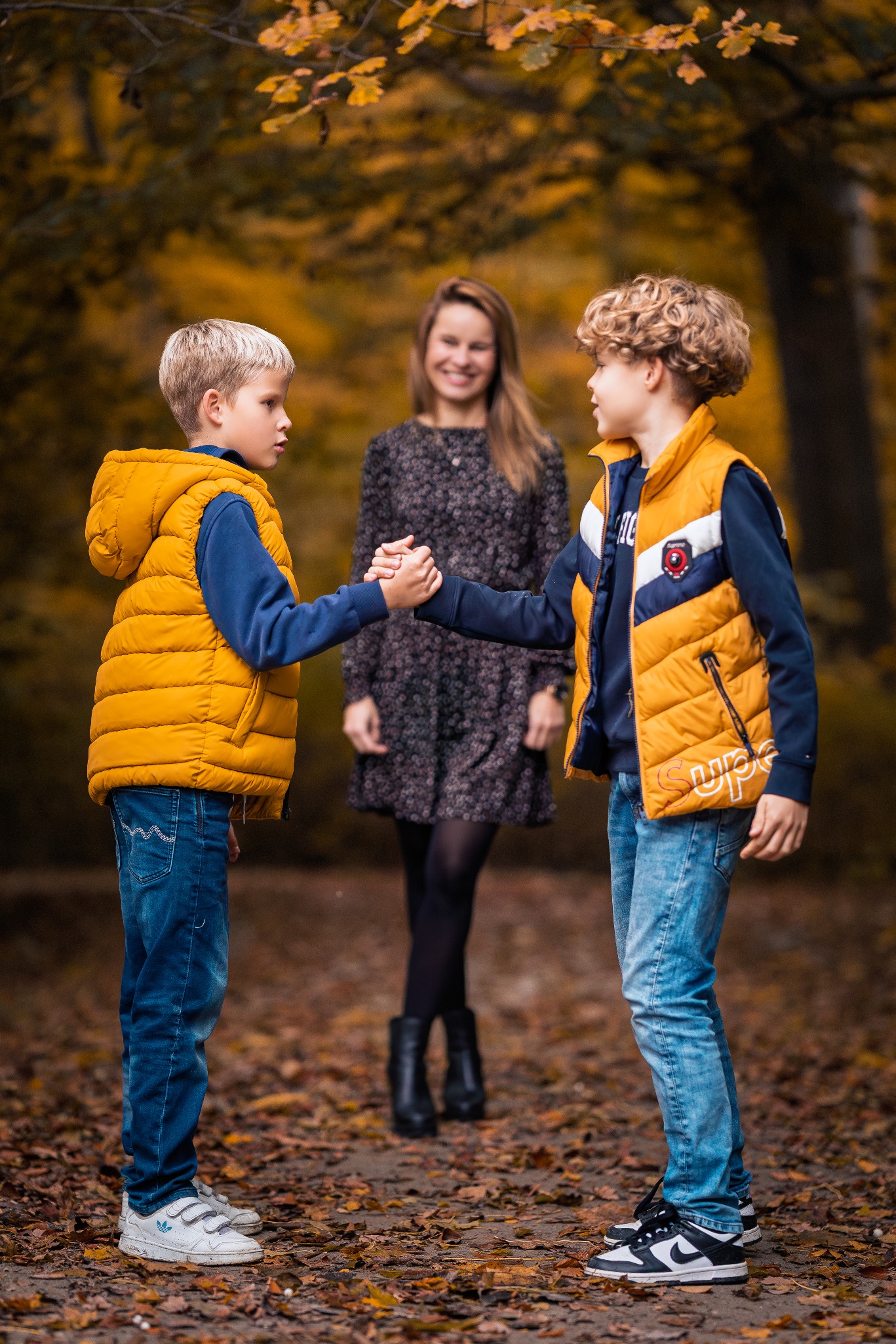 Jesienna sesja rodzinna: Dwóch chłopców w żółtych kamizelkach ściska dłonie na tle uśmiechniętej kobiety w parku, opadłe liście, ciepłe światło.