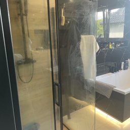 Czysty Dom - Zabrudzona kabina prysznicowa z czarnymi ramami i marmurowym wykończeniem. Widoczne ślady po wodzie i mydle na szklanych drzwiach. W tle wanna i półka z kosmetykami.