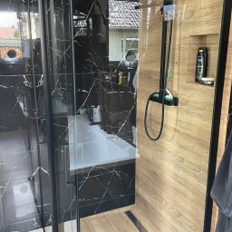 Czysty Dom - Nowoczesna kabina prysznicowa z czarnymi marmurkowymi płytkami i drewnopodobnymi panelami, czarna armatura, widok na wannę i okno.