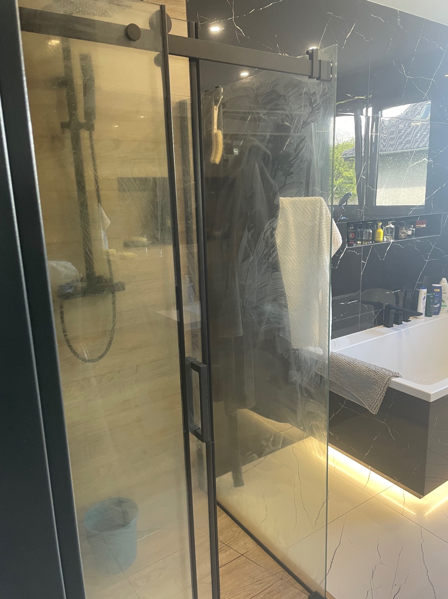Zabrudzona kabina prysznicowa z czarnymi ramami i marmurowym wykończeniem. Widoczne ślady po wodzie i mydle na szklanych drzwiach. W tle wanna i półka z kosmetykami.