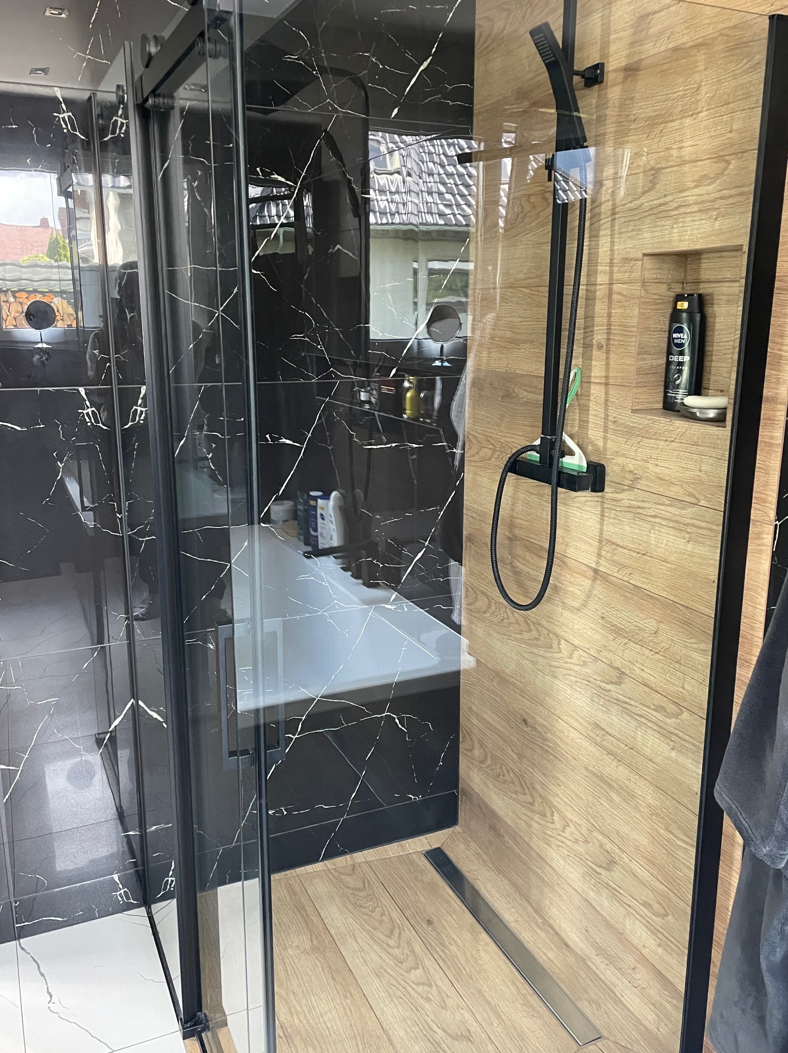 Nowoczesna kabina prysznicowa z czarnymi marmurkowymi płytkami i drewnopodobnymi panelami, czarna armatura, widok na wannę i okno.