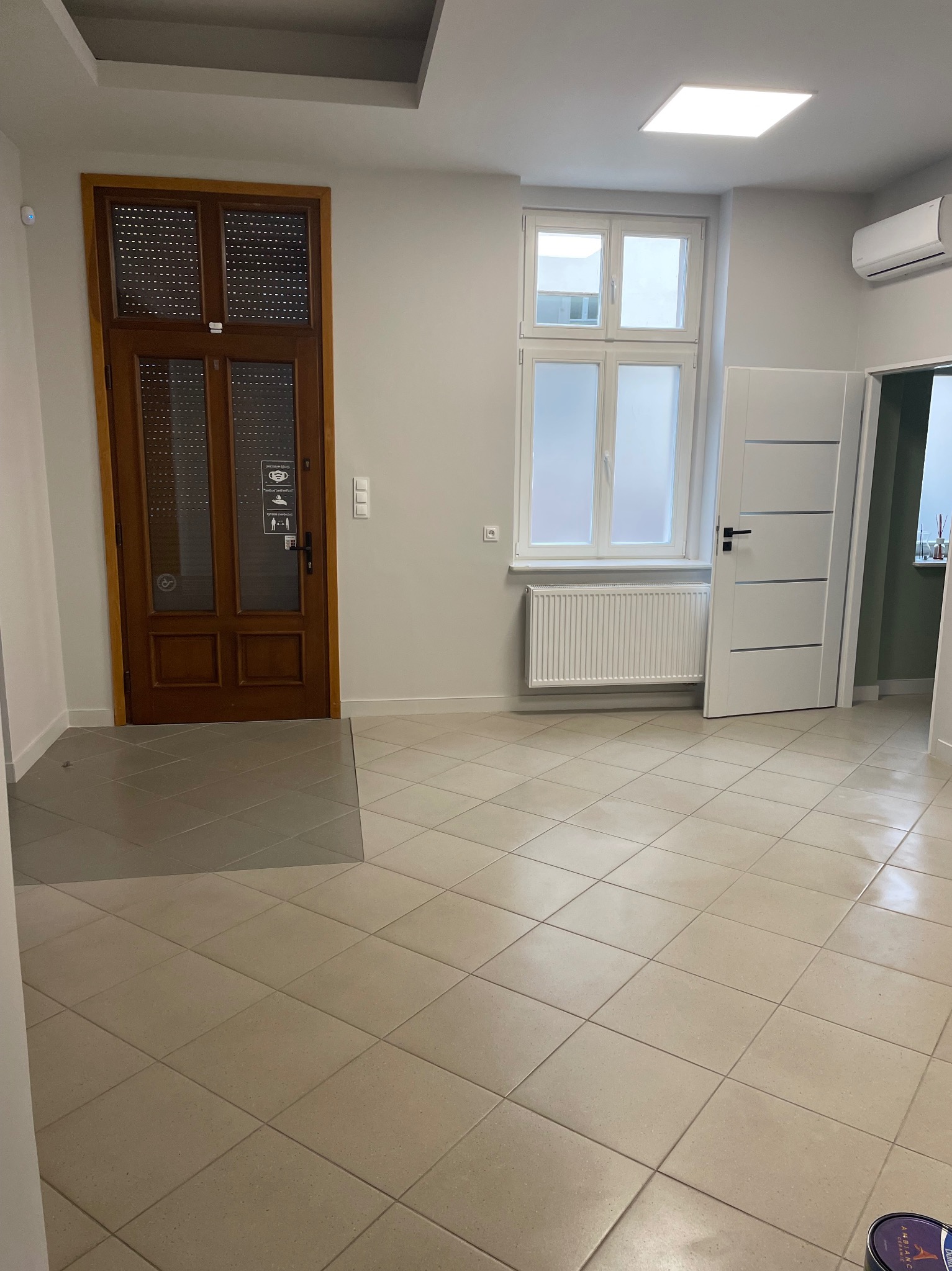 Jasne, przestronne wnętrze po sprzątaniu: beżowe płytki podłogowe, białe ściany, okno z grzejnikiem, drewniane drzwi z informacjami i białe drzwi do kolejnego pomieszczenia.