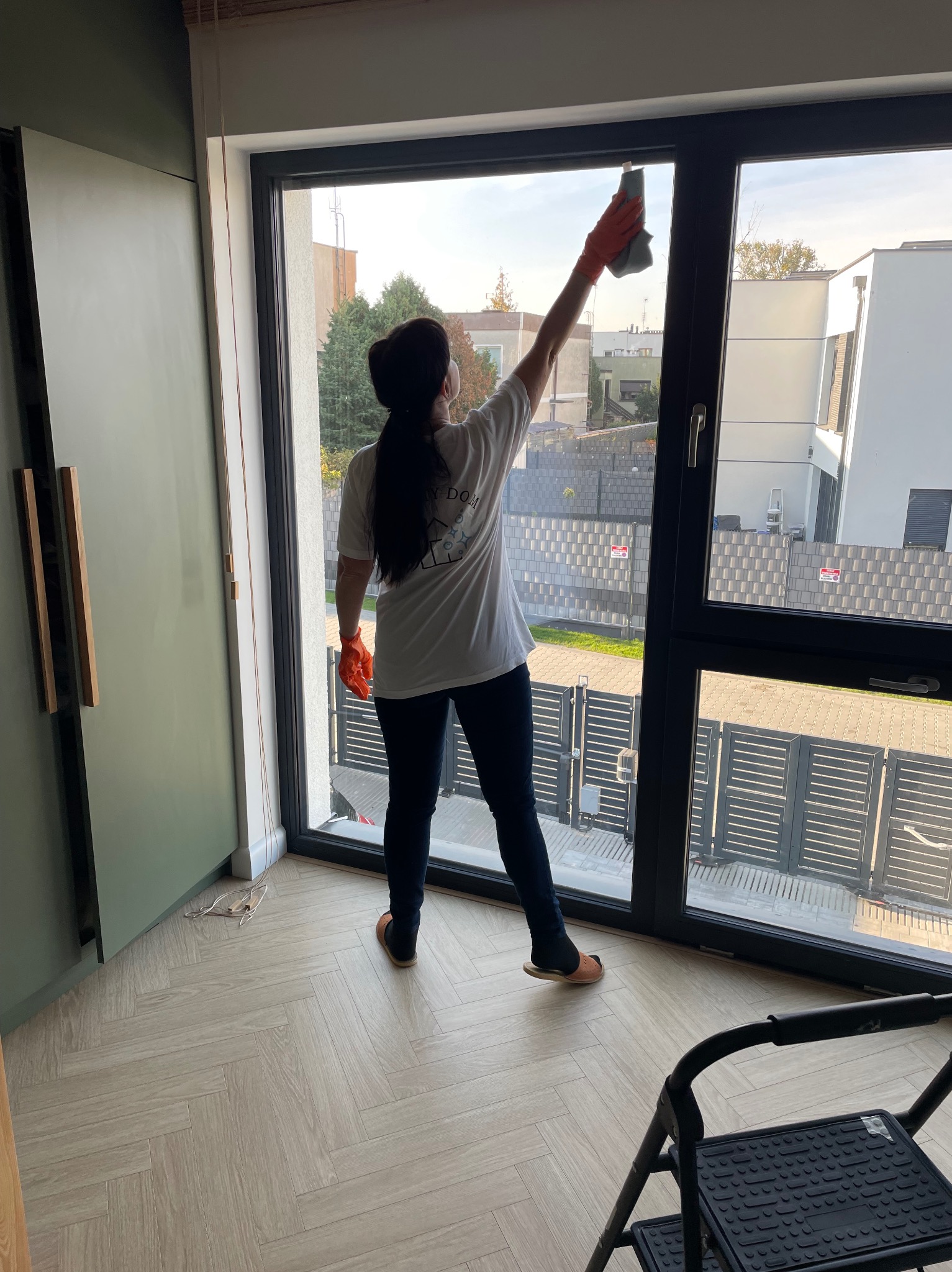 Kobieta w rękawicach ochronnych czyści duże okno w nowoczesnym wnętrzu. Widoczna drabina, podłoga w jodełkę i zabudowa meblowa w kolorze oliwkowym.