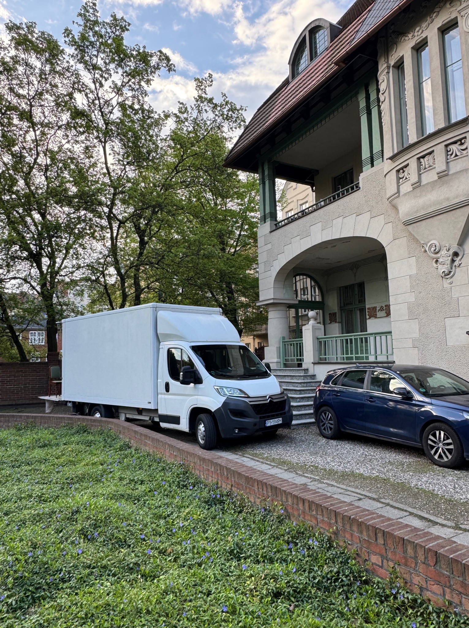 Biały samochód dostawczy zaparkowany przed secesyjną kamienicą, obok stoi niebieski samochód osobowy. Widoczne drzewa i roślinność.